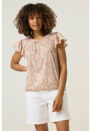 Nainen kiharat hiukset, yllään lyhythihainen kukallinen paita ja valkoiset, käänteillä varustetut shortsit yksiväristä taustaa vasten.