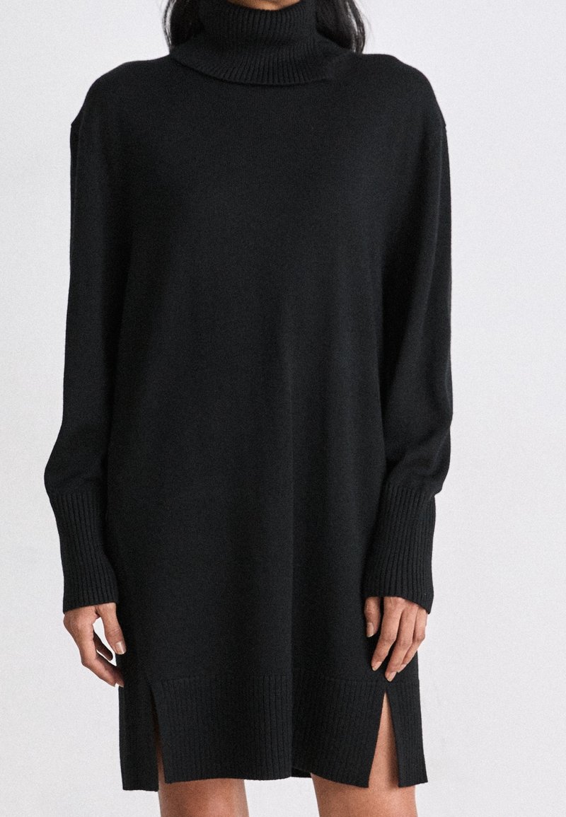 Žena v černé dlouhé svetr s rolákem, který má žebrované manžety, lem a boční rozparky, stojící na jednobarevném pozadí.