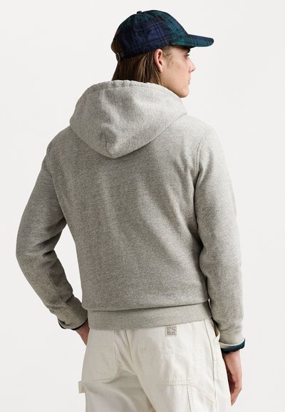 Sweat à capuche gris avec une capuche, des poignets et un ourlet côtelés. Porté avec un pantalon de couleur claire et une casquette à motif. Design minimaliste et tissu texturé.