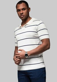 Vit kortärmad polo med marinblå horisontella ränder, med en ribbad krage och en figurnära design. Bärs med mörka jeans och en klocka.