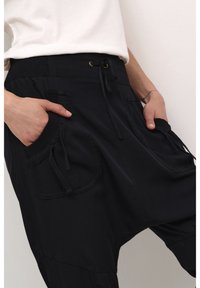 Personne portant un pantalon noir ample avec de grandes poches latérales et une taille à cordon, mains dans les poches sur un fond uni.