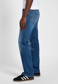 Blue denim jeans with a straight cut, featuring back pockets and subtle stitching. Worn with black sneakers featuring white stripes.