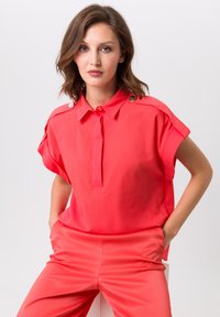 Koraalkleurig shirt met korte mouwen en een kraag, met een knoopsluiting en metalen accentknopen op de schouders, gecombineerd met bijpassende koraalkleurige broek.