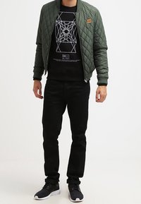 Veste bomber matelassée olive avec des poignets en bord-côte, associée à un sweat-shirt graphique noir et un pantalon noir. Les chaussures sont des baskets en mesh noires.