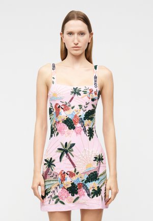 Γυναίκα που φοράει αμάνικο ροζ ανοιχτό φόρεμα με πολύχρωμους κεντημένους παπαγάλους, φοίνικες, λουλούδια και σχέδιο ηλιοβασιλέματος, στέκεται μπροστά σε απλό φόντο.