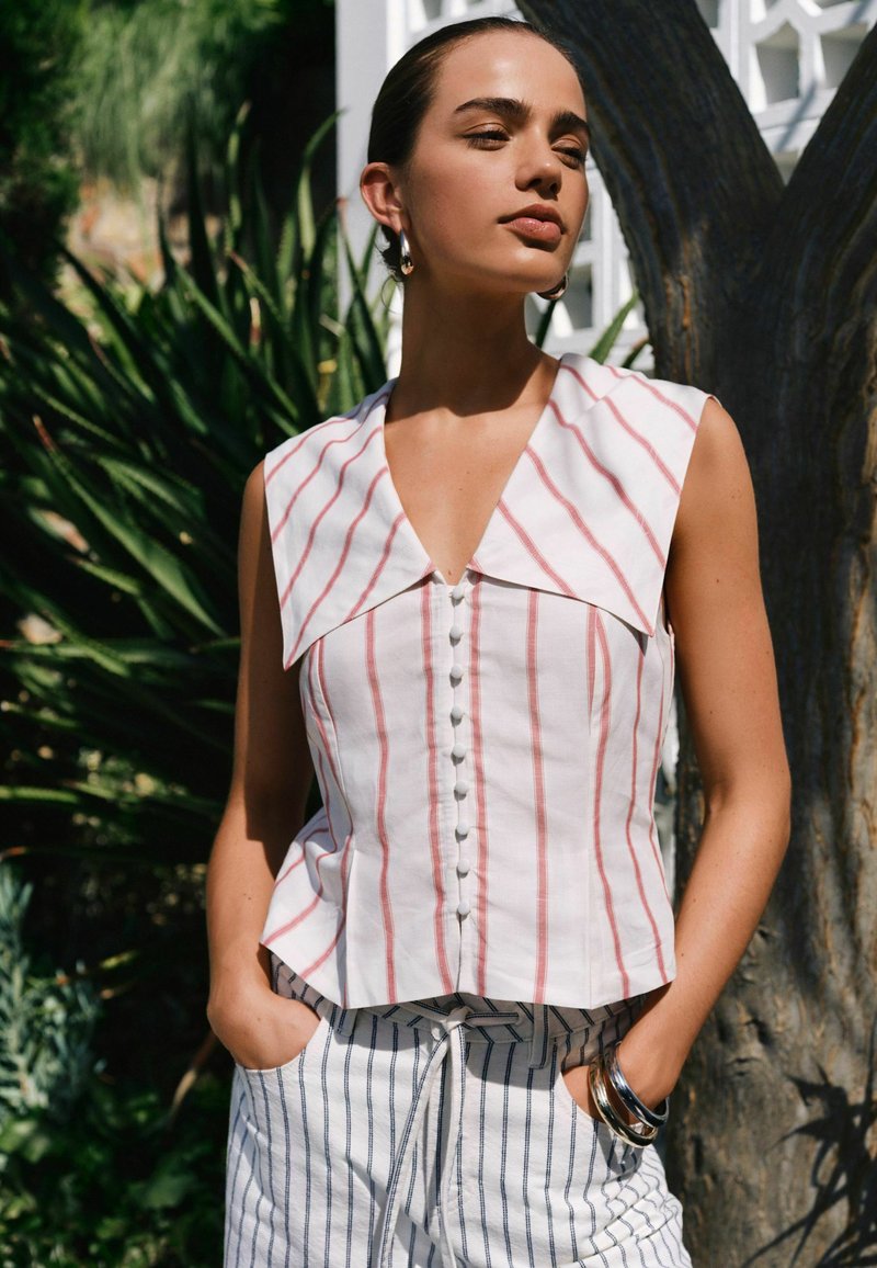 Femme debout à l'extérieur, portant un haut sans manches blanc à rayures rouges et un pantalon blanc à rayures noires, les mains dans les poches, à côté d'un arbre.