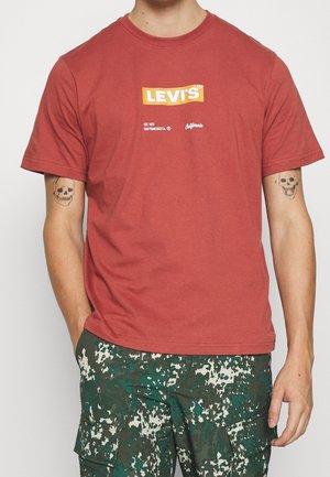 Person trägt ein rostfarbenes Levi's T-Shirt und grün-weiße Tarn-Cargohosen, die Hände in den Taschen, mit Totenkopf-Tattoos auf beiden Unterarmen.
