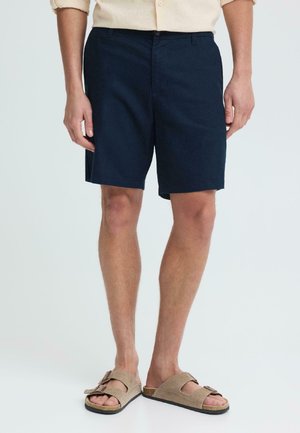Mand iført marineblå shorts, beige skjorte og brune ruskindssandaler, stående mod en ensartet hvid baggrund.