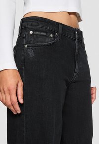 Black denim jeans featuring slight distressing, classic five-pocket design, silver hardware, and a Calvin Klein label on the front pocket.