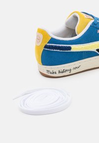 Modré semišové tenisky s žlutými akcenty, bílými šňůrkami a texturovanými detaily. Na boku se nachází podpis: "Udělej historii teď."