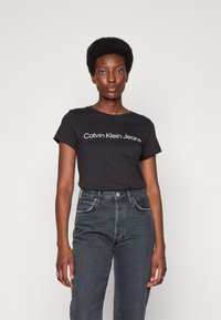 Černé bavlněné tričko s bílým textem "Calvin Klein Jeans", kombinované s tmavými džíny s vysokým pasem. Krátké rukávy a kulatý výstřih.