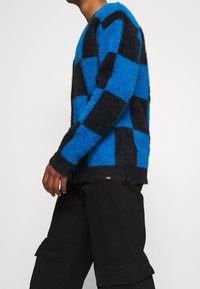 Lämmin neulepusero, jossa on sininen ja musta ruutukuosi, rento leikkaus ja ribbaattiset hihansuut. Yhdistetty mustiin cargo-housuihin.