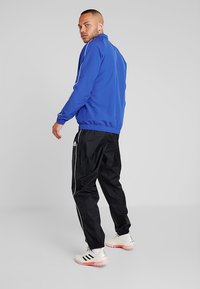 Blaue Trainingsjacke mit weißen Streifen, schwarze Trainingshose und weiße Sneaker mit roten Akzenten, von hinten vor einem grauen Hintergrund betrachtet.