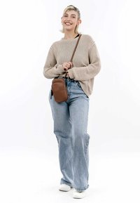 Borsa a tracolla marrone con texture liscia, forma rettangolare e dettaglio con cerniera, indossata sopra un maglione di maglia beige e jeans a gamba larga blu.