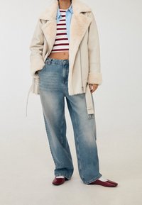 Béžový kabát s podšívkou z beránka, červený a bílý pruhovaný crop top, světle modrá košile na knoflíky, široké džíny se vybledlým vzhledem a červené boty na blokovém podpatku.