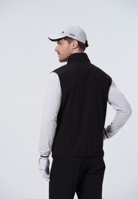 Zwarte mouwloze vest over een lichtgrijs langetermijnshirt, gedragen met een grijze pet, met een hoge kraag en elastische manchetten.