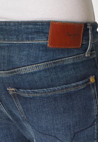 Tył niebieskich jeansów denim z brązową skórzaną metką Pepe Jeans London oraz przeszywaną tylną kieszenią z małym metalowym guzikiem.