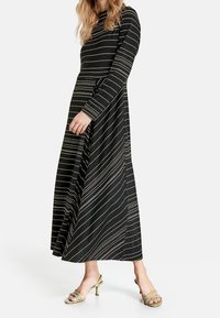 Robe noire à manches longues avec des rayures horizontales dorées et une silhouette en A. Associée à des sandales à talons vertes. Texture de tissu lisse.