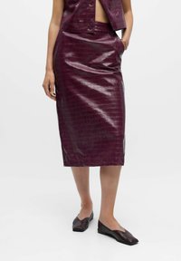 Bordeaux faux leren kokerrok met krokodillenstructuur, tot de knie en met een zijzak. Gepaard met zwarte platte schoenen.