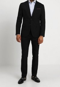 Svart kostym med skräddarsydd design, enkelknäppning på kavajen, vit och blå randig skjorta, svart bälte och blanka loafers. Röd accent på fickan.