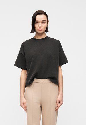 Giovane con capelli corti e scuri che indossa una maglietta nera larga e pantaloni beige a vita alta, in piedi davanti a uno sfondo chiaro e uniforme.