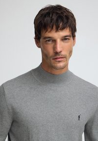 Pull gris en maille avec un col montant, arborant un petit logo noir brodé sur la poitrine. Texture douce et coupe ajustée.