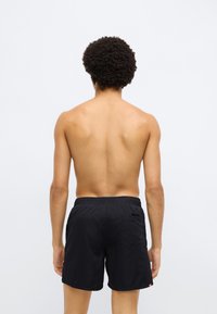 Mustad ujumispüksid elastse vöökohaga ja tagataskuga, millel on sile tekstuur ja lihtne disain, nähtud tagantpoolt.