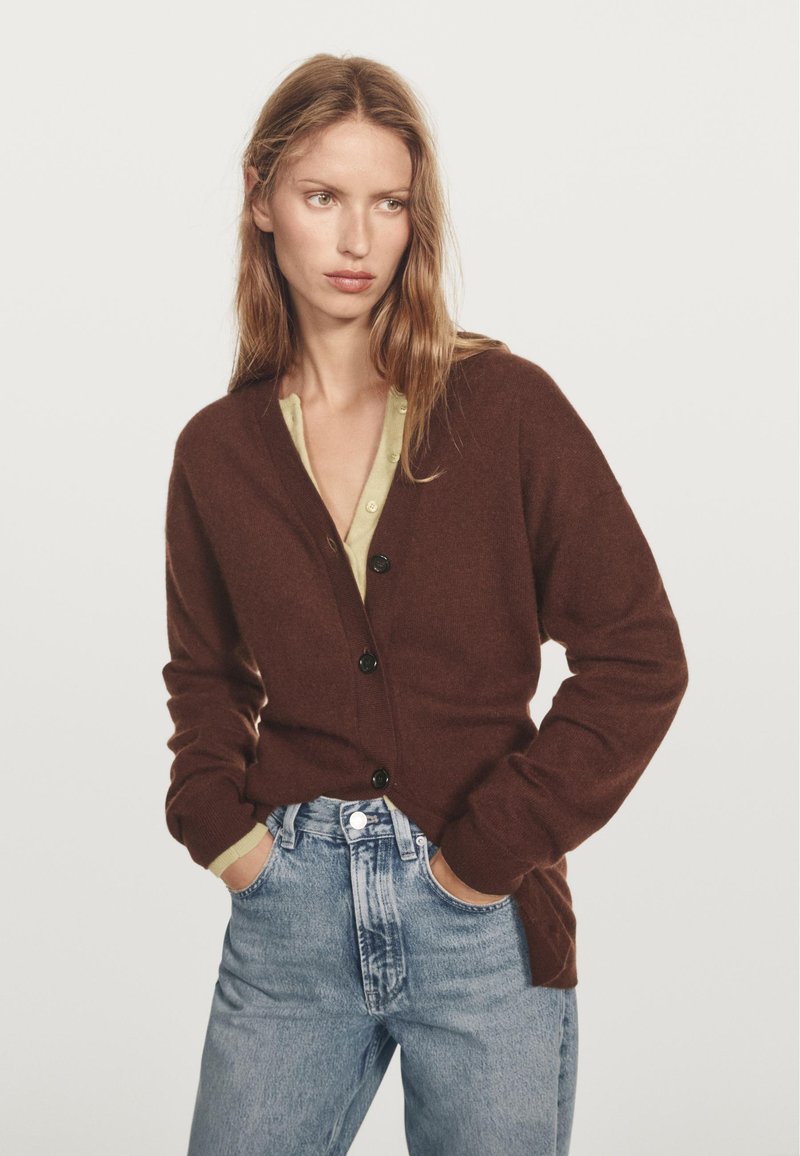 Hnědý cardigan s černými knoflíky, dlouhými rukávy a límcem do V. Nosený přes světle zelený top a kombinovaný s džíny s vysokým pasem.