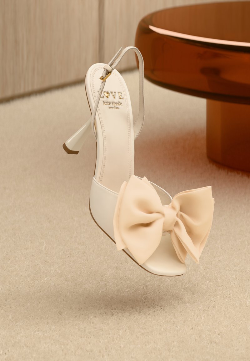 Bílé sandály na vysokém podpatku s velkou měkkou broskvovou mašlí vpředu, čirými popruhy a béžovou stélkou s detailním zlatým logem.