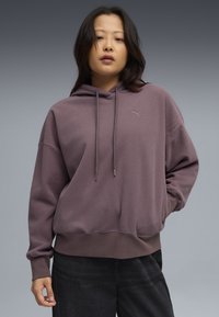Przesadny fioletowy hoodie z teksturowanej tkaniny, wyposażony w kieszeń kangurzą, kaptur ze sznurkiem oraz małe logo na klatce piersiowej.