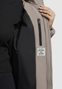 Lightweight, beige outer jacket with a smooth texture. Interior features black lining and a zip pocket, labeled “A warm hug made out of cloth.”