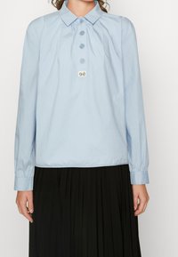 Camicia a maniche lunghe di colore azzurro chiaro con colletto, cinque bottoni e un piccolo logo su un'etichetta rettangolare, indossata sopra una gonna nera plissettata.