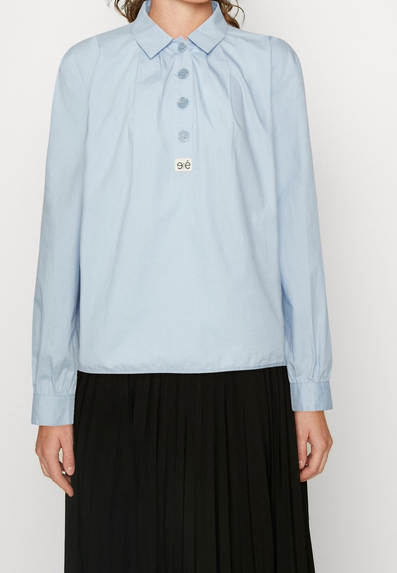 Camicia a maniche lunghe di colore azzurro chiaro con colletto, cinque bottoni e un piccolo logo su un'etichetta rettangolare, indossata sopra una gonna nera plissettata.