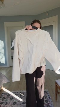 Pulover crem din tricot de tip cablu, cu mâneci largi și lejere și umeri căzuți, având modele texturate și un croi relaxat, agățat pe umeraș.