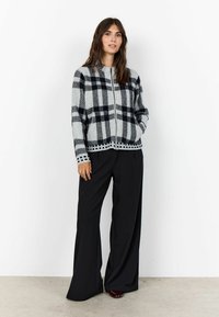 Sweter kardigan w szaro-czarną kratę z zamkiem z przodu i okrągłym dekoltem; zestawiony z czarnymi spodniami o szerokich nogawkach i burgundowymi butami.