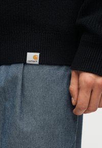 Main reposant à côté d'un pantalon en denim bleu, portant un pull noir à côtes avec une petite étiquette blanche du logo Carhartt à l'ourlet.