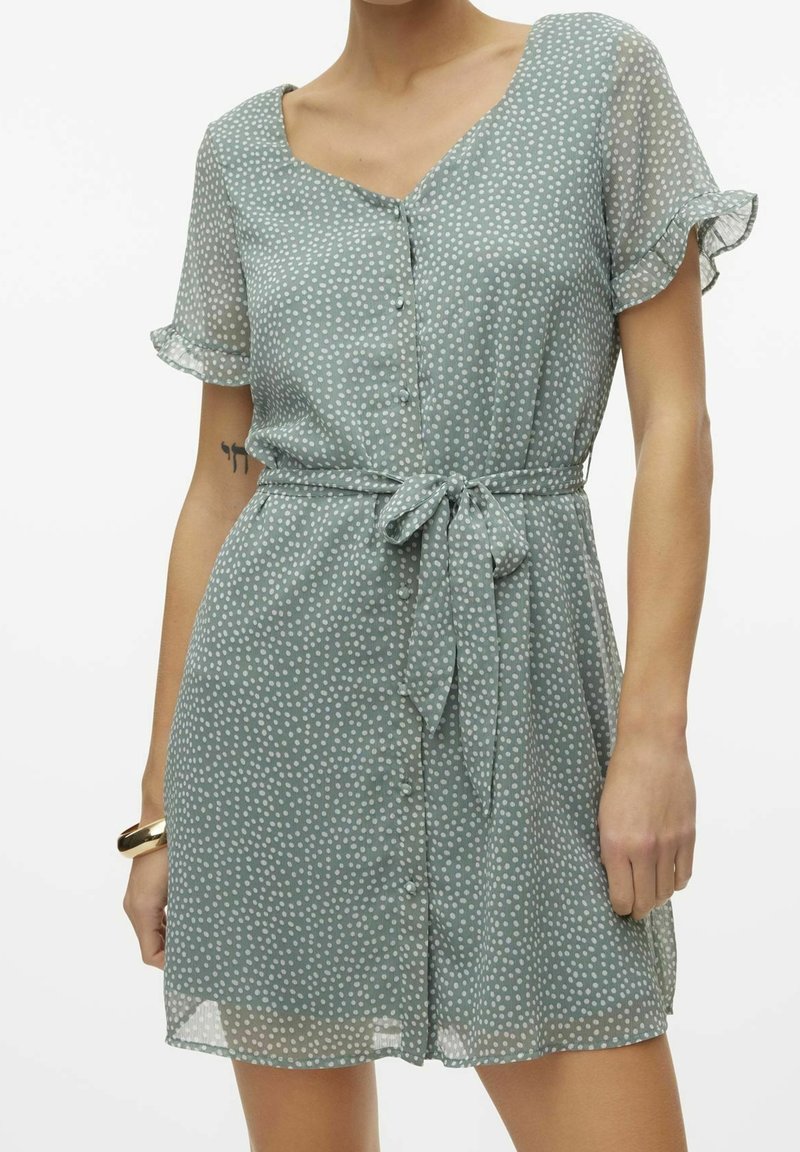 Grünes Polka-Dot-Kleid mit kurzen, flatternden Ärmeln, V-Ausschnitt, durchgehender Knopfleiste und einem Stoffgürtel. Leichtes, fließendes Material mit weicher Textur.