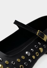Μαύρο παπούτσι από συνθετικό υλικό με στρογγυλεμένη μύτη, διαθέτει χρυσά και ασημένια καρφιά κατά μήκος της πλευράς και λεπτό λουράκι αστραγάλου με αγκράφα.