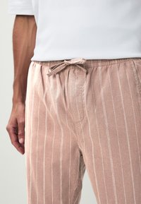 Lyserøde bukser med striber, elastik i taljen, en justerbar snøre og blød tekstur. Har hvide lodrette striber for ekstra detalje.