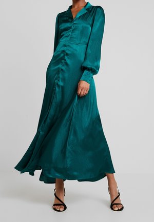 Kvinde iført teal satin langærmet kjole med knapper kombineret med sorte remsandaletter med høje hæle mod en grå baggrund.