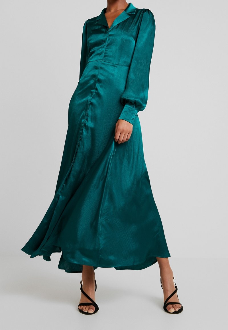 Femme portant une robe boutonnée en satin vert-bleu à manches longues, associée à des sandales noires à talons hauts et à lanières, sur fond gris.