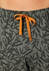 Close-up of waist shows gray pants with black leaf pattern and orange drawstring tied in a bow, worn by a person with exposed skin.