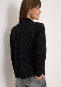 Zwarte lange mouwen jas met een gestructureerd luipaardprint patroon. Voorzien van een kraag en casual pasvorm, gecombineerd met grijze jeans.
