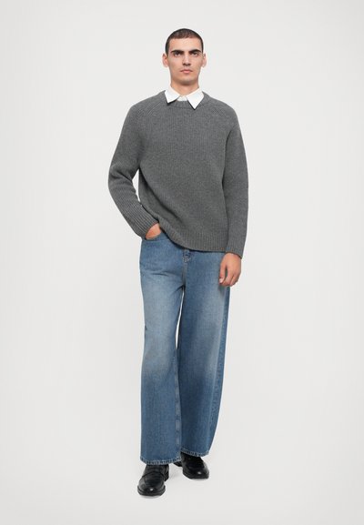 Pull en tricot gris avec un col rond, associé à un jean bleu clair à jambes larges. Le mannequin se tient avec une main dans la poche et des chaussures noires.