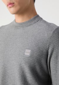 Homme portant un pull gris texturé avec un petit écusson carré du logo BOSS sur la poitrine gauche, sur un fond clair uni.