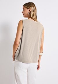 Top sin mangas beige con una textura suave, escote redondeado y corte holgado, combinado con pantalones anchos blancos. Diseño simple y versátil.