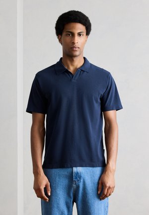 Man standing wearing a dark blue short-sleeve polo shirt and light blue jeans against a plain light background.