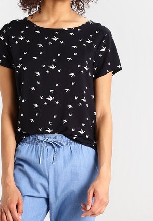 Femme portant un t-shirt noir à manches courtes avec des imprimés d’oiseaux blancs et un pantalon bleu clair à cordon, sur un fond clair uni.