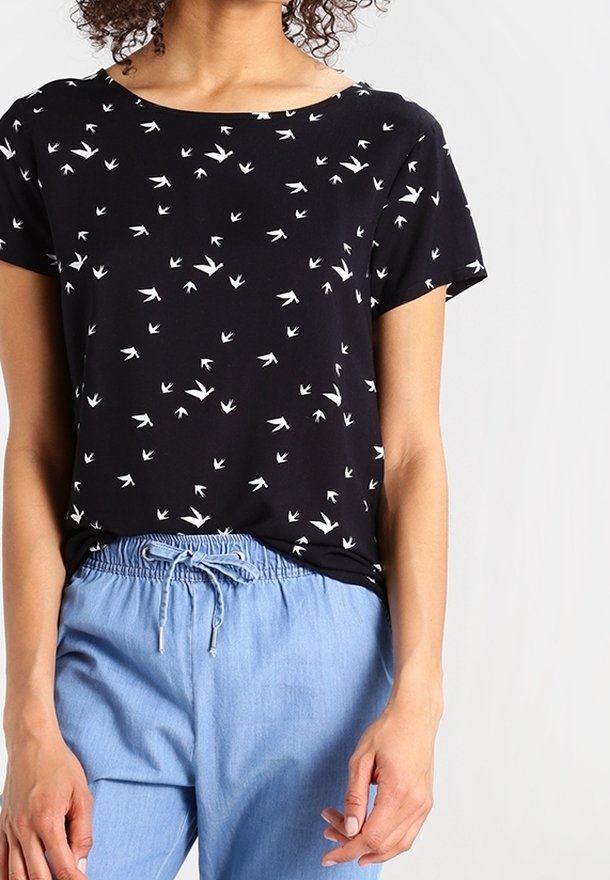 Femme portant un t-shirt noir à manches courtes avec des imprimés d’oiseaux blancs et un pantalon bleu clair à cordon, sur un fond clair uni.