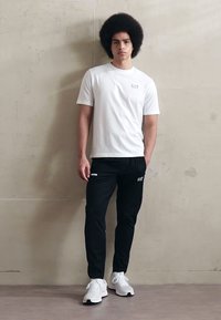 Fehér, rövid ujjú póló kis logóval; fekete nadrág logóval; fehér sportcipő. Simára varrott anyag, testhezálló stílusban.